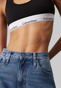 Bralette negro con banda blanca de logo de Calvin Klein, combinada con jeans de mezclilla azul de cintura alta que presentan un botón y diseño clásico de cinco bolsillos.