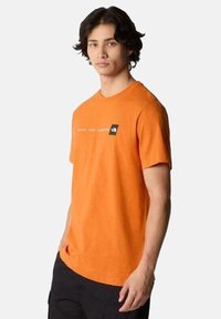 Camiseta de algodón naranja con cuello redondo, mangas cortas, que presenta un pequeño logo negro y texto en el pecho. Textura suave.