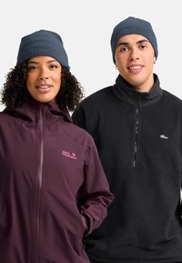 Due persone che indossano berretti navy e giacche Jack Wolfskin, una con giacca a vento viola e l'altra con pullover in pile nero, sorridono di fronte.
