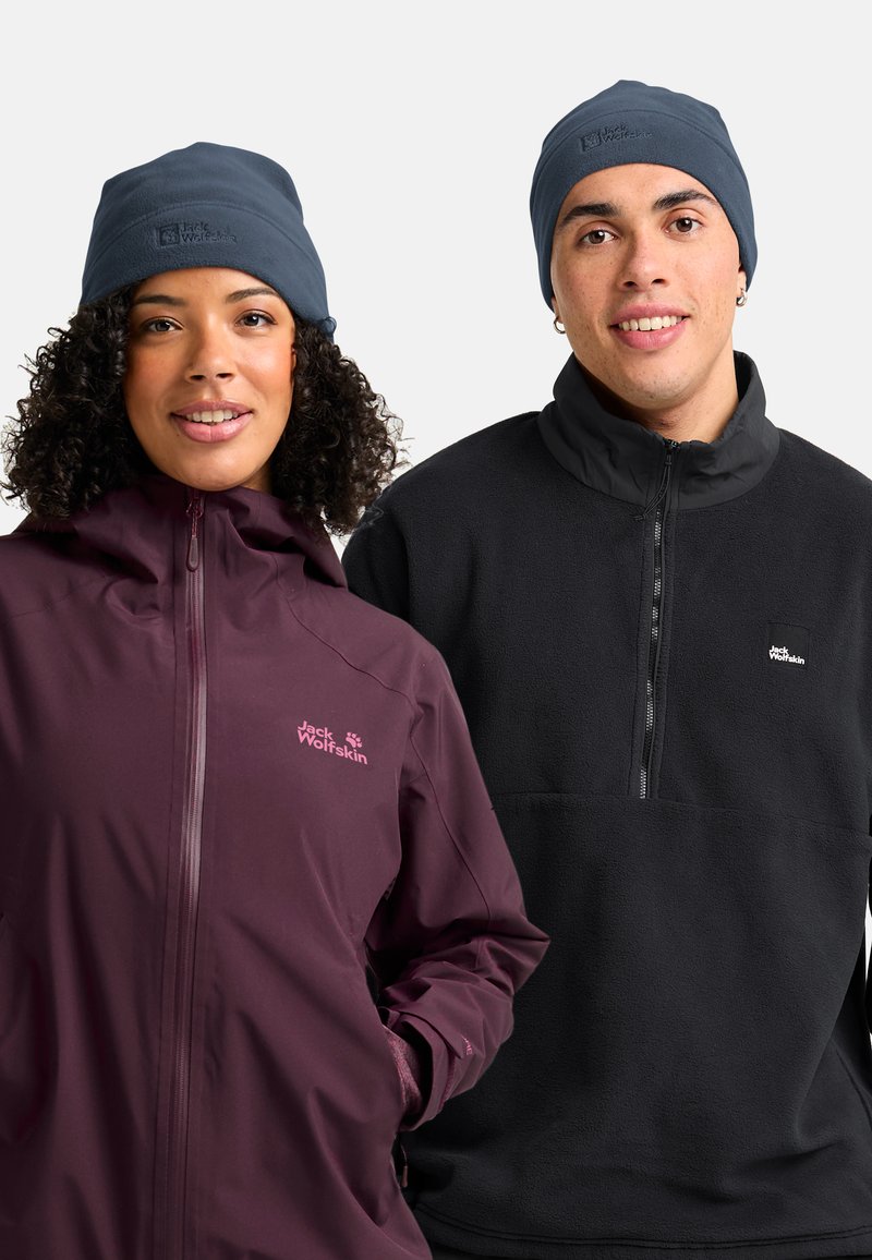 Due persone che indossano berretti navy e giacche Jack Wolfskin, una con giacca a vento viola e l'altra con pullover in pile nero, sorridono di fronte.