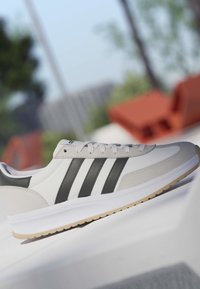 Biały sneaker z czarnymi paskami i beżową podeszwą, prezentowany w plenerze z rozmytymi drzewami i pomarańczowymi siedzeniami w tle.