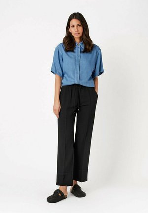 Woman standing with hands in pockets wearing a short-sleeve blue shirt, black drawstring pants, and black slip-on shoes against white background.