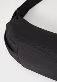 Juodas „Guess" krepšys su lygiu, tekstūriniu paviršiumi, turintis kreivą formą ir iškilnų logotipo detalę. Reguliuojama dirželis universalumui.