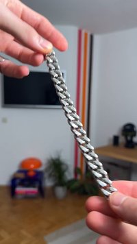 Masyívny strieborný náramok z prepojených očiek, ktorý odráža svetlo. Hladký kovový povrch s leštenou textúrou, držený medzi prstami.