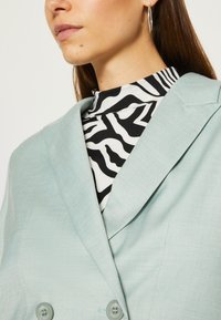 Világoskék, kétsoros blézer texturált felülettel, fekete-fehér zebra mintájú, magas nyakú felsővel párosítva, fülbevalókkal.