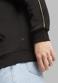 Felpa nera con polsini a costine, caratterizzata da una striscia dorata lungo il braccio. Il tessuto è morbido, con una vestibilità slim, abbinata a jeans blu scoloriti.