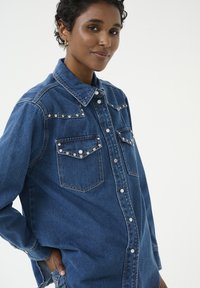 Denim krekls ar garām piedurknēm, poga ar snap tipa aizdari un divām priekšējām kabatām. Iespēju rotājumi un klasiskā zilā nokrāsa.