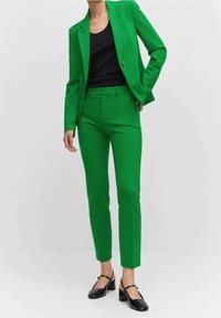 Zielona marynarka i spodnie w gładkim materiale, połączone z czarną bluzką i czarnymi butami na kwadratowym obcasie.