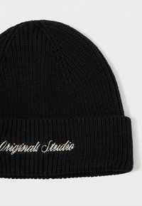 Sort ribbet strikbeanie med foldet kant, broderet med hvid kursiv tekst "Originals Studio" på hvid baggrund.