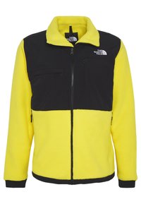 Žlutá fleecová bunda s černým panelem na hrudi, zapínáním na zip vpředu, bočními kapsami a logem The North Face na levé straně hrudi a uvnitř límce.