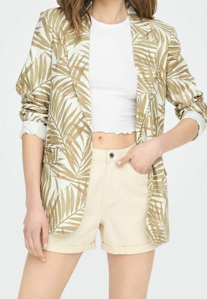 Femme portant un blazer beige et blanc à motif de feuilles, un haut court blanc côtelé et un short beige clair, la main dans la poche.