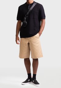 Musta teksturoitu poolopaita, jossa on tasku, yhdistettynä beigeihin polvipituisiin shortseihin. Asun täydentävät mustat liukuslip-on kengät ja pieni olkalaukku.