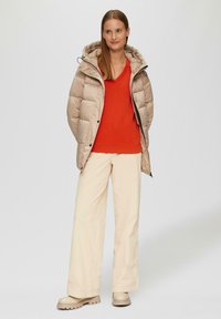 Beżowa kurtka puchowa z kapturem, zestawiona z żywym czerwonym topem i kremowymi spodniami o szerokich nogawkach. Masywne beżowe buty dodają tekstury.