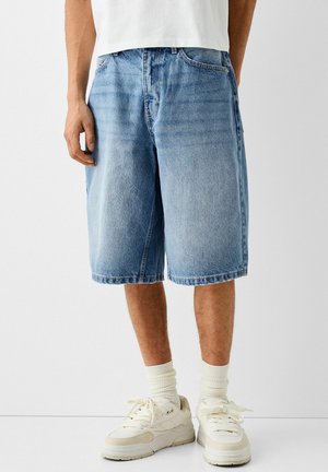 Person trägt kniehohe, lockere blaue Jeansshorts, weiße Socken und weiße Turnschuhe und steht vor einem einfachen hellen Hintergrund.