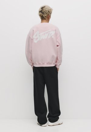 Osoba o krótkich blond włosach, ubrana w oversize'ową różową bluzę z białym graficznym napisem na plecach, czarne spodnie i białe trampki.