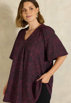 Mujer que lleva un top floral suelto de color morado oscuro con mangas cortas y anchas y escote en V, pendientes de aro dorados y pantalones negros, posando con una mano en el bolsillo.