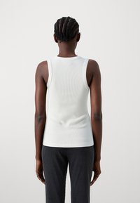 Bílý žebrovaný tílko s přiléhavým střihem, bez rukávů a kulatým výstřihem, v kombinaci s tmavými strukturovanými kalhotami.