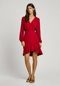 Robe rouge à manches longues, avec un ourlet froncé et un tissu texturé. Présente une taille cintrée et un décolleté en V. Portée avec des sandales marron.