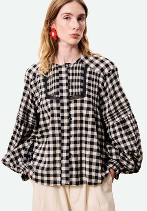 Femme portant un chemisier à carreaux noir et blanc avec des manches bouffantes et des boucles d’oreilles rouges en forme de fleur, debout les mains dans les poches.