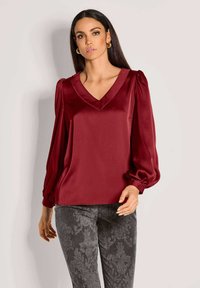 Burgunderrote Satinbluse mit V-Ausschnitt, Puffärmeln und glatter Textur. Kombiniert mit dunklen gemusterten Hosen mit einem Damastdesign.