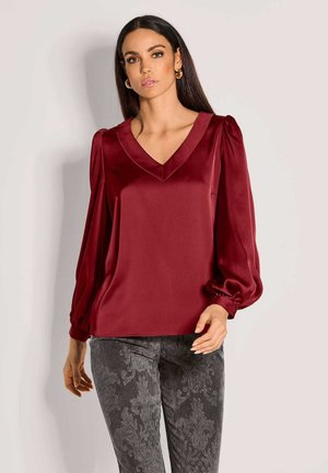 Burgunderrote Satinbluse mit V-Ausschnitt, Puffärmeln und glatter Textur. Kombiniert mit dunklen gemusterten Hosen mit einem Damastdesign.