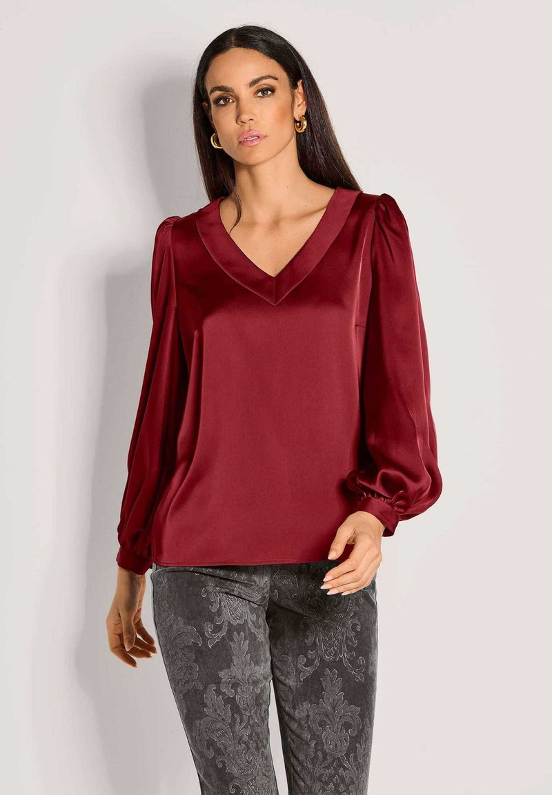 Burgunderrote Satinbluse mit V-Ausschnitt, Puffärmeln und glatter Textur. Kombiniert mit dunklen gemusterten Hosen mit einem Damastdesign.
