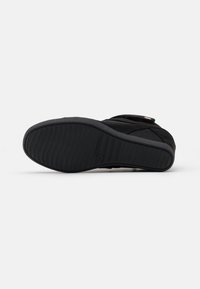 Svart atletisk sko med texturerat överdel och ett mönstrat gummisula. Har en kardborreband och en diskret logotypdetalj nära hälen.