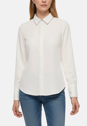 Blouse blanche à manches longues avec un col pointu, présentant un détail décoratif en chaîne sur le col et la patte de boutonnage, texture lisse.