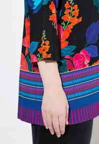 Kukkainen tunika, jossa on musta tausta ja kirkkaat punaiset, siniset ja vihreät värit. Helma on varustettu geometrisella kuvioinnilla.