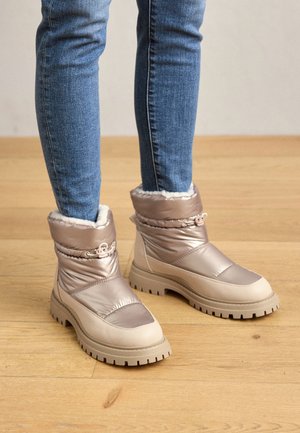 Person iført beige polstrede vinterstøvler med tykke såler, stående på et trægulv, iført blå skinny jeans.