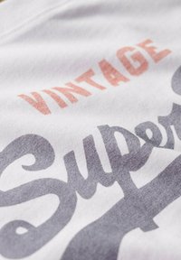 Weißes Stoff-T-Shirt mit einem Vintage-Design, das "VINTAGE" in hellem Rosa und "Super" in dunkelgrauer Schreibschrift zeigt.