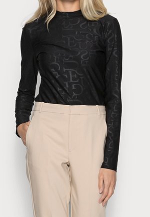 Femme portant un haut noir à manches longues avec des motifs subtils de lettres, associé à un pantalon beige taille haute, main dans la poche côté gauche.
