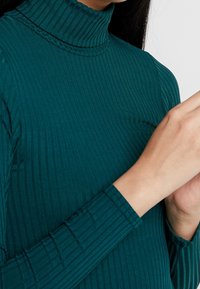 Teal ribbattuotupaita pitkillä hihoilla; siinä on tiukka muotoilu ja hienovarainen tekstuuri. Lähikuva näyttää kaula-aukkoalueen ja hihojen yksityiskohdat.