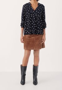 Femme portant un chemisier bleu marine à pois à manches longues, une jupe en velours côtelé marron, et des bottes en cuir noires mi-mollet, debout sur un fond uni.