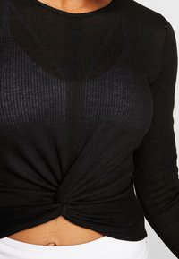 Svart ribbat långärmad topp med en tvistad knutdetalj vid midjan. Mjuk textur med en åtsittande design, rund halsringning och minimala sömmar.
