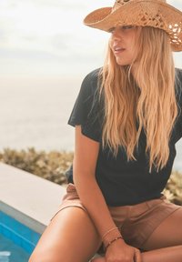 Čierne tričko, svetlohnedé šortky a široký slamenný klobúk. Mäkké, vlnité blond vlasy, so náramkom na zápästí. Okolie bazéna.