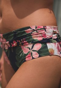 Gėlėtos bikini kelnaitės, kuriose dera rožinės gėlės ir žali lapai ant tamsios spalvos fono, su surinkta audinio dalimi juosmens srityje.