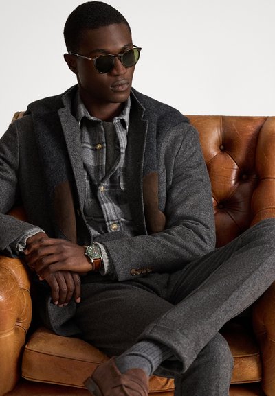 Mężczyzna w ciemnych okularach przeciwsłonecznych, szarej warstwowej odzieży, z brązowym skórzanym zegarkiem i brązowymi butami, siedzący ze skrzyżowanymi nogami na brązowej skórzanej kanapie.