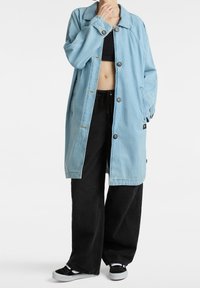 Giacca oversized in denim blu chiaro con colletto, chiusura a bottoni, tasche laterali e maniche morbide, indossata sopra pantaloni neri a gamba larga.