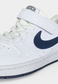 Λευκό αθλητικό παπούτσι Nike με σκούρο μπλε λογότυπο swoosh, λευκά κορδόνια και μια λωρίδα με βέλκρο στο πάνω μέρος, σε κοντινή λήψη.