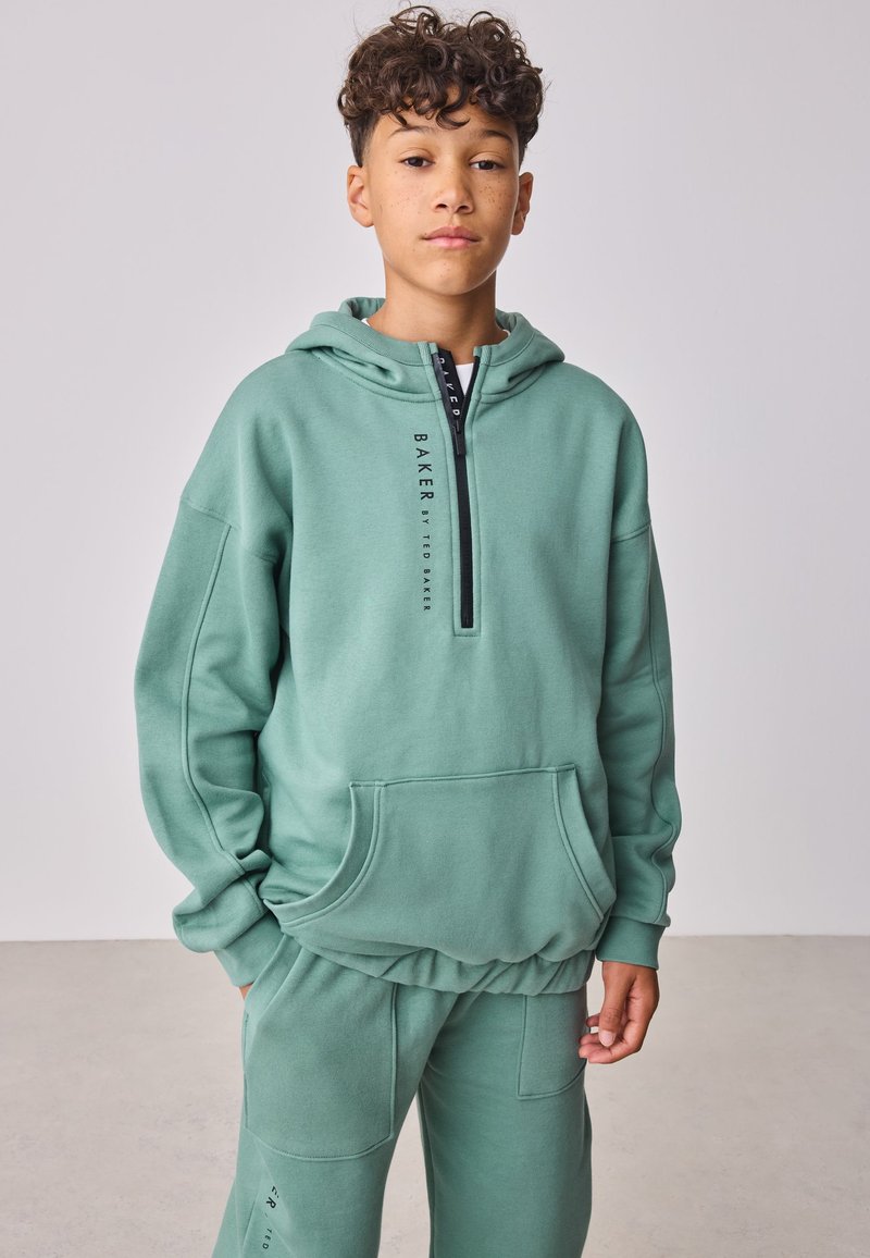 Poiss lokkis juustega, kes kannab helerohelist kapuutsiga pusa ja sobivaid dressipükse, seisab siseruumides tavalise heleda tausta ees.