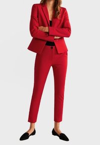 Rød skræddersyet blazer og matchende tapered bukser i et glat materiale, med en enkelt knaplukning og et slankt design. Sorte flade sko fuldender looket.