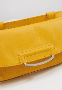 Sac à main en cuir jaune avec coutures visibles et détail de poignée en métal argenté sur un fond clair uni.