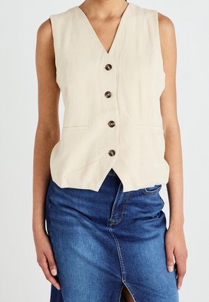 Veste beige sans manches avec col en V, cinq boutons marron, et deux poches devant, associée à une jupe en denim. Texture lisse, coupe décontractée.