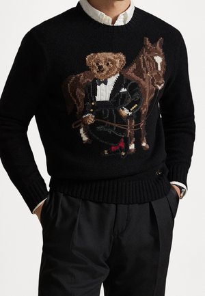 Schwarzer Pullover mit einem gestrickten Design eines Bären im Smoking, der die Zügel neben einem braunen Pferd hält, über einem weißen Hemd getragen.