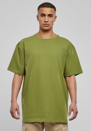 Giovane uomo con capelli corti, indossa una maglietta oversize verde oliva e pantaloni cargo beige, in piedi contro uno sfondo grigio chiaro uniforme.