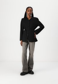 Blazer noir structuré avec un col en V, fermeture à bouton et taille ceinturée, associé à un jean gris évasé et des chaussures à plateforme noires.