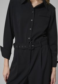 Fekete, hosszú ujjú kezeslábas, galléros nyakkal, gombos elülső résszel, mellzsebbel és széles övvel, amely téglalap alakú csattal rendelkezik. Sima szövet állag.