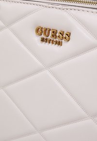 Borsa a tracolla in pelle chiara con motivo a rombi trapuntato. Logo "GUESS" in oro e dettaglio in rilievo "EST 1981". Chiusura con zip.