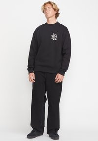 Sudadera negra con cuello redondo y diseño gráfico en el lado izquierdo del pecho, combinada con pantalones negros de corte holgado y zapatillas negras.